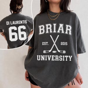 May include: Dark grey t-shirt with white text and graphics. The front displays "BRIAR UNIVERSITY" with crossed hockey sticks and a puck. The back shows "DI LAURENTIS" and the number "66".
