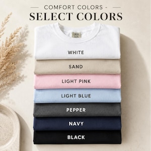 May include: A stack of seven folded t-shirts in various colors labeled Comfort Colors Select Colors. The colors from top to bottom are white, sand, light pink, light blue, pepper, navy, and black. Each shirt is neatly folded and stacked vertically.