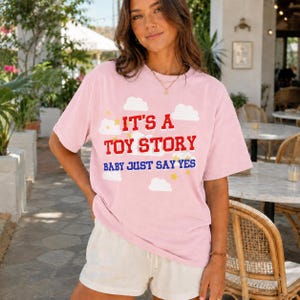 May include: A light pink short-sleeved t-shirt featuring a graphic print of white clouds and yellow stars. The text reads IT'S A TOY STORY in red block letters and BABY JUST SAY YES in blue letters. The shirt has a crew neckline and loose fit.