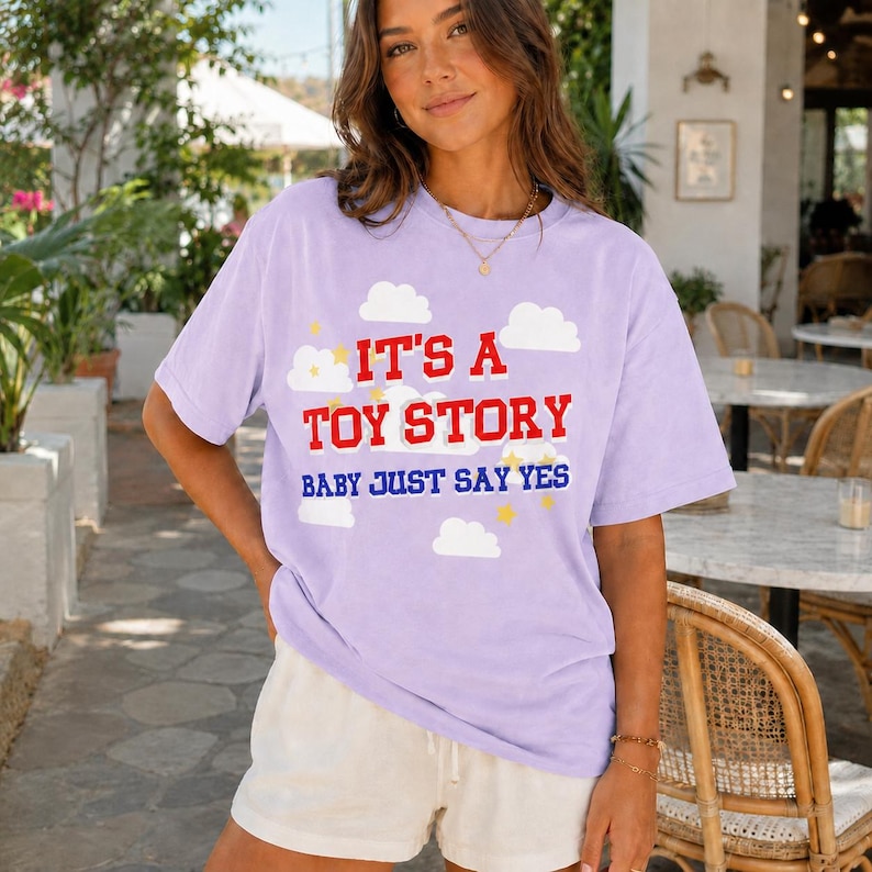 May include: A light purple short-sleeve t-shirt featuring a graphic print of white clouds and yellow stars. The text reads IT'S A TOY STORY in red block letters and BABY JUST SAY YES in blue letters. The shirt is paired with white shorts.