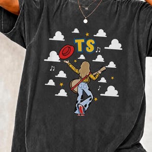 Peut inclure: Un t-shirt gris anthracite avec un motif repr&eacute;sentant une personne aux cheveux longs portant une chemise &agrave; carreaux jaune et un jean pattes d'eph &agrave; motifs de vache, tenant une guitare et un chapeau rouge. Le design inclut des nuages blancs.