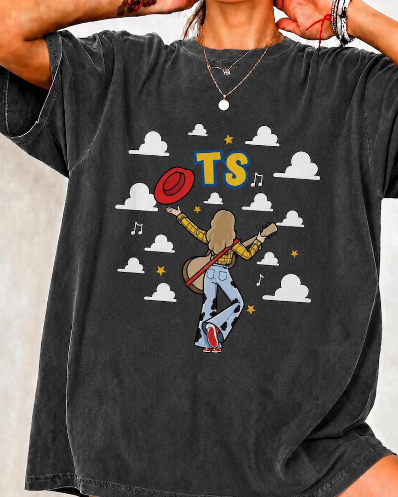 Peut inclure: Un t-shirt gris anthracite avec un motif repr&eacute;sentant une personne aux cheveux longs portant une chemise &agrave; carreaux jaune et un jean pattes d'eph &agrave; motifs de vache, tenant une guitare et un chapeau rouge. Le design inclut des nuages blancs.
