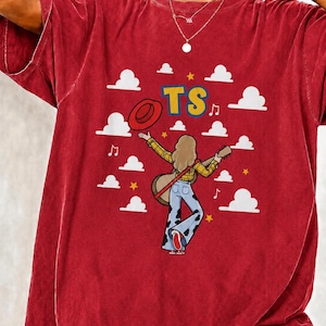Peut inclure: Un t-shirt rouge &agrave; manches courtes avec un motif repr&eacute;sentant une personne aux cheveux longs portant une chemise &agrave; carreaux jaune et un jean pattes d'eph &agrave; taches de vache, tenant une guitare et un chapeau rouge, avec des nuages et le texte TS.