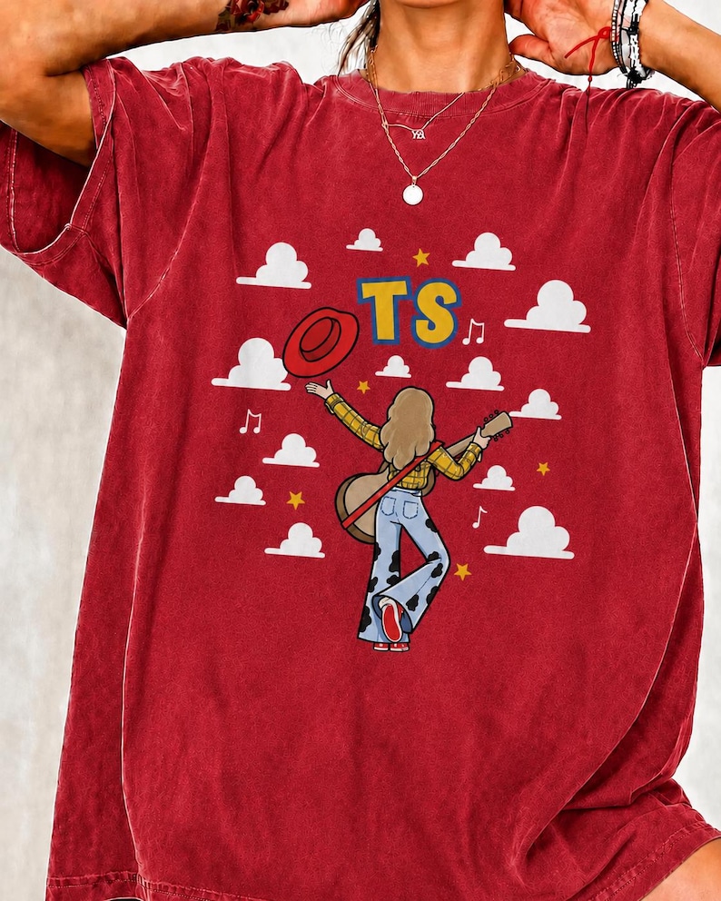 Peut inclure: Un t-shirt rouge &agrave; manches courtes avec un motif repr&eacute;sentant une personne aux cheveux longs portant une chemise &agrave; carreaux jaune et un jean pattes d'eph &agrave; taches de vache, tenant une guitare et un chapeau rouge, avec des nuages et le texte TS.