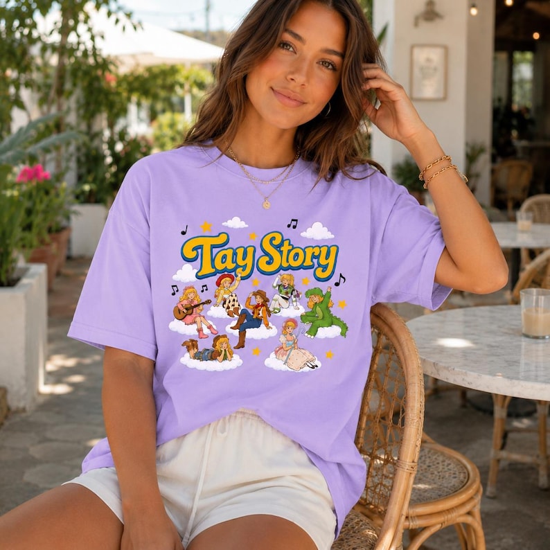 May include: A light purple short-sleeve t-shirt featuring a graphic print with the text Tay Story in yellow letters. The design includes various cartoon characters sitting on white clouds, surrounded by small yellow stars and black musical notes.