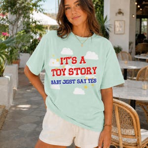 May include: A light mint green short-sleeve t-shirt featuring a graphic print of white clouds and yellow stars. The text reads IT'S A TOY STORY in red block letters and BABY JUST SAY YES in blue letters centered on the front of the garment.