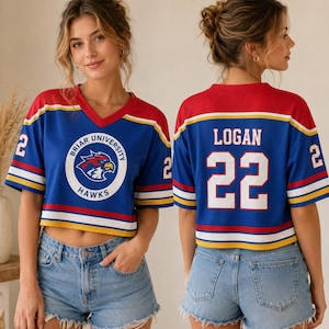 Op de afbeelding: Blauw, rood en wit cropped jersey met de tekst "Briar University Hawks" en het nummer "22". De jersey heeft een V-hals en wordt gedragen met een spijkerbroek. De jersey heeft de naam "Logan" op de rug.
