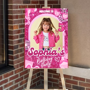 Op de afbeelding: Een roze en glitterig verjaardagsfeestbord op een ezel. Het bord zegt "Welkom bij 5 Sophia's Verjaardagsfeest" met een foto van een jong meisje in een roze jasje. Decoratieve elementen zijn harten, discoballen en bloemen.