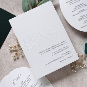 May include: Elegant white wedding invitation with embossed text reading "Claire & Samuel." Includes details for the ceremony, RSVP information, and a welcome party. A green envelope and ring box with gold rings are also visible.