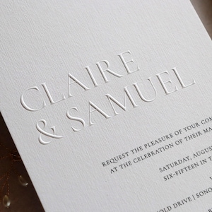 May include: Elegant white wedding invitation with embossed text reading "CLAIRE & SAMUEL." The invitation includes details for the celebration, such as the date, time, and location. The paper has a textured finish.