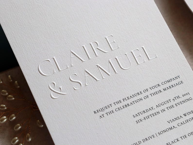 May include: Elegant white wedding invitation with embossed text reading "CLAIRE & SAMUEL." The invitation includes details for the celebration, such as the date, time, and location. The paper has a textured finish.