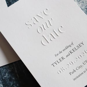 May include: Close-up of a white embossed wedding invitation. The text "save our date" is prominently displayed. Additional text includes the names TYLER and KELSEY, the date 08.29.2026, and location Park City, UT. The invitation is for a wedding.