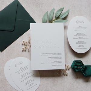 May include: Elegant wedding stationery set featuring a white invitation with embossed text, matching details cards, and a dark green envelope. Two rings rest in a green velvet ring box, with floral accents.