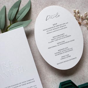 May include: Elegant white wedding invitation suite. Includes a rectangular card with embossed names and a rounded details card with event information. Greenery and a pearl accent add a touch of sophistication.