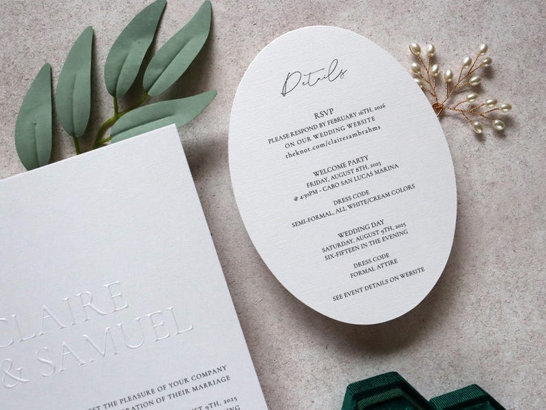 May include: Elegant white wedding invitation suite. Includes a rectangular card with embossed names and a rounded details card with event information. Greenery and a pearl accent add a touch of sophistication.