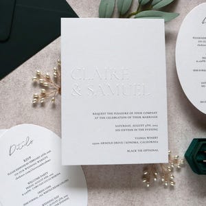 May include: Elegant wedding invitation suite featuring embossed text "Claire & Samuel" on a white card. Includes details cards and a dark green velvet ring box. The invitation includes the date, time, and location of the wedding.