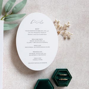May include: Wedding invitation suite featuring a white oval card with event details, including RSVP information and dress codes. Two gold rings rest in a green velvet box. A sprig of pearls and greenery are also present.