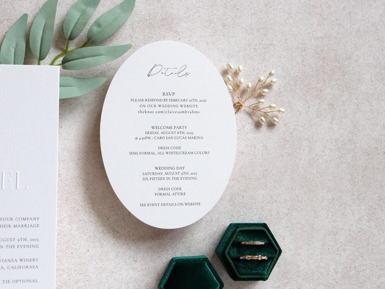 May include: Wedding invitation suite featuring a white oval card with event details, including RSVP information and dress codes. Two gold rings rest in a green velvet box. A sprig of pearls and greenery are also present.