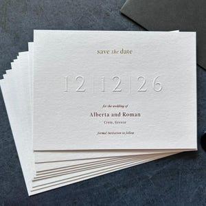 Op de afbeelding: Een stapel witte trouwkaarten met reliëfcijfers en tekst. De kaarten tonen "save the date" en "12 12 26". De tekst hieronder luidt "for the wedding of Alberta and Roman, Crete, Greece".