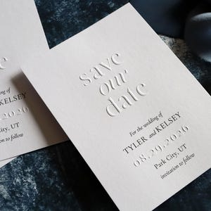 May include: Off-white save the date cards with embossed lettering reading save our date, for the wedding of Tyler and Kelsey, 08.29.2026, Park City, UT, invitation to follow. The cards feature a textured paper surface and dark printed text.