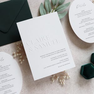 May include: Elegant wedding invitation suite featuring embossed text with the names "Claire & Samuel". Includes a dark green envelope, details cards, and a velvet ring box. The invitation details the wedding date and location.