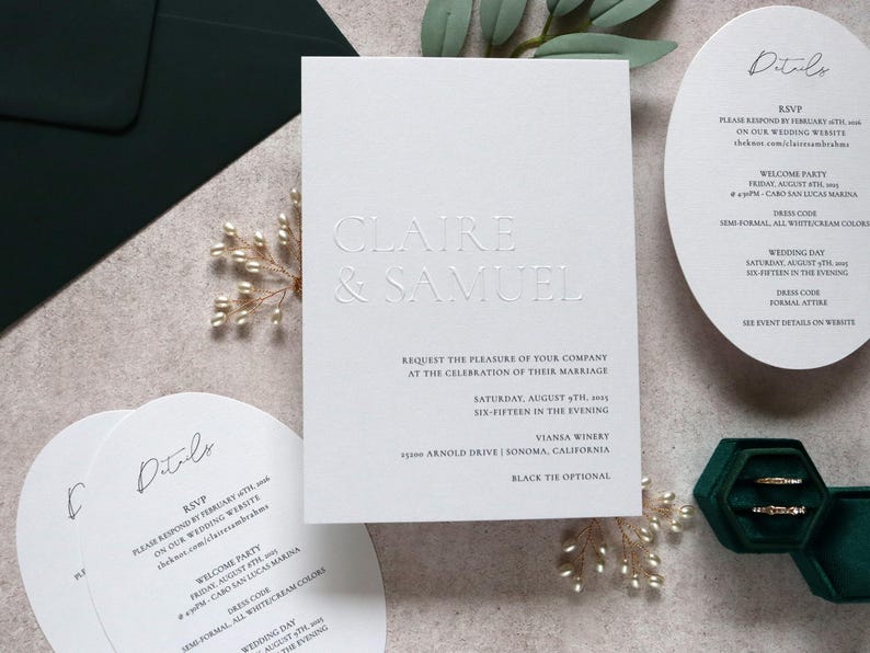 May include: Elegant white wedding invitations with embossed text, including the names "Claire & Samuel." Details cards and a ring box with gold rings are also present. The invitations are on a neutral surface with greenery.