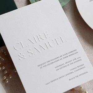 May include: A white wedding invitation with embossed text reading "CLAIRE & SAMUEL." The invitation includes details for the ceremony: date, time, and location. The text is in a simple, elegant font.