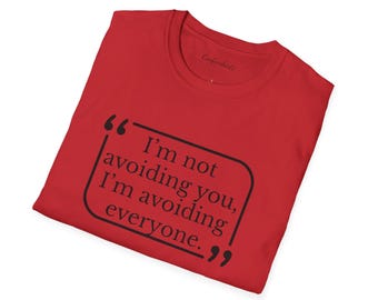 May include: A red t-shirt featuring the quote "I'm not avoiding you, I'm avoiding everyone" in a black speech bubble. The shirt is made of a soft material, suitable for casual wear.