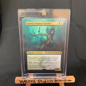 May include: A trading card featuring Zegana, Utopian Speaker, a Merfolk Wizard. The card is in a protective case, displayed on a small wooden easel with "Weerd Studios" written on it. The card has a teal and green color scheme.