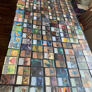 May include: A large collection of trading cards, primarily Magic: The Gathering, spread out on a dark surface. The cards feature various fantasy art and text, with a variety of colors and designs. The cards are arranged in rows and columns.