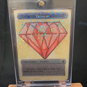 May include: A custom-made Treasure token card featuring a large red diamond illustration. The card is encased in a protective sleeve and displayed on a small wooden easel. The text "TREASURE" is at the top, with "Weerd Studios 3" written on the easel.