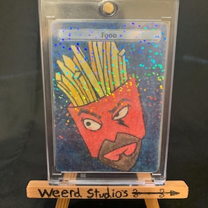 May include: A trading card featuring a cartoon character designed as a red french fry container with a face and fries. The word "Food" is at the top. The card is in a clear protective case, displayed on a small wooden easel. The easel has the text "Weerd Studios".