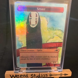 May include: A holographic trading card featuring a watercolor illustration of a character named Spirit. The card is encased in a protective sleeve and displayed on a small wooden easel. The card's text reads "Token Creature - Spirit" and "Menace".