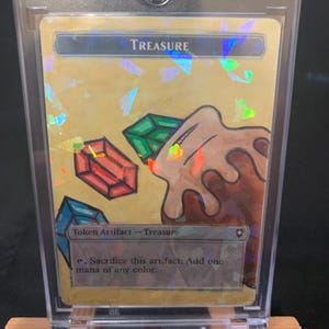 May include: A holographic "Treasure" card in a protective case. The card features a gold background with colorful geometric shapes and illustrations of gems and a brown blob. The text "Token Artifact - Treasure" is visible.