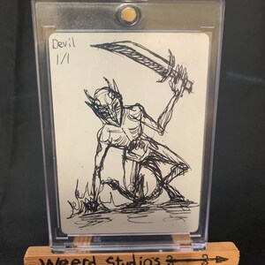 May include: A sketch card featuring a black ink drawing of a devil figure holding a sword. The card is labeled "Devil 1/1". The card is displayed in a clear protective case on a small wooden easel with "Weerd Studios" printed on it.