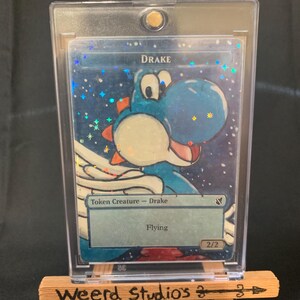 May include: A blue and white Drake token creature card with a cartoon illustration of a Yoshi-like character. The card is in a clear protective case and displayed on a small wooden easel. The text "Drake" and "Flying" are visible.