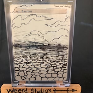 May include: A trading card in a protective case, featuring a black and white landscape illustration with the text "Ash Barrens." The card is displayed on a small wooden easel with the text "Weerd Studios."