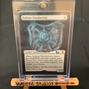 May include: A trading card titled "Solemn Simulacrum" in a protective case, displayed on a small wooden easel. The card features a blue and white illustration and text. The easel is branded with "Weerd Studios 3".