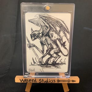 May include: A black and white sketch of a devil with large wings, horns, and a skeletal appearance. The artwork is displayed in a clear protective case on a small wooden easel. The text "Devil 1/1" is visible.