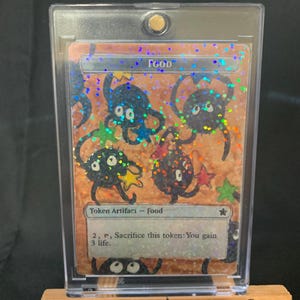 May include: A clear plastic card holder displaying a "Food" token artifact card. The card features cartoonish black creatures with star-shaped appendages on a speckled orange background. The text "Weerd Studio's" is visible on the wooden stand.