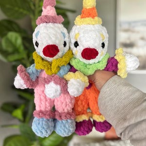 May include: Two handmade crochet clown dolls made from soft, plush yarn. The left doll features a pink hat, a mustard yellow collar, and light blue pants. The right doll has a yellow and orange hat, a lime green collar, and orange pants.