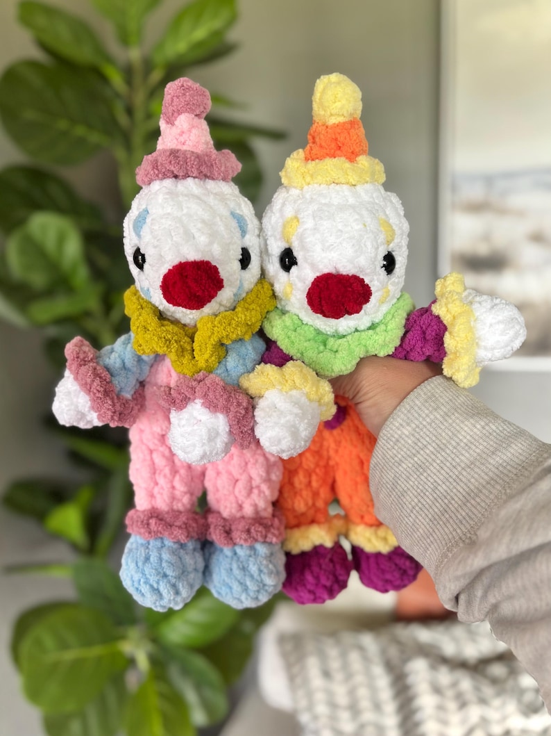 May include: Two handmade crochet clown dolls made from soft, plush yarn. The left doll features a pink hat, a mustard yellow collar, and light blue pants. The right doll has a yellow and orange hat, a lime green collar, and orange pants.