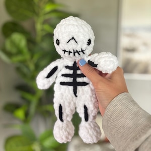 May include: A white plush skeleton doll made of soft yarn with black embroidered details on the face, rib cage, and limbs. The skeleton features a rounded head, stitched mouth, and simple black lines representing bones on its torso and arms.