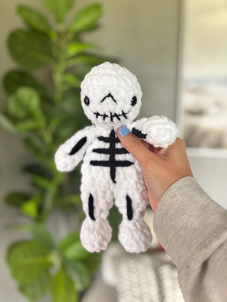 May include: A white plush skeleton doll made of soft yarn with black embroidered details on the face, rib cage, and limbs. The skeleton features a rounded head, stitched mouth, and simple black lines representing bones on its torso and arms.