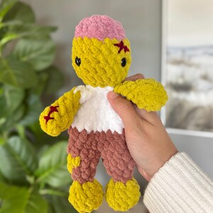 May include: A handmade crochet plush doll featuring a yellow head with a pink top, black safety eyes, and red stitched details. The doll has a white torso, brown legs, and yellow feet, crafted from soft, chunky yarn with a textured, knotted appearance.