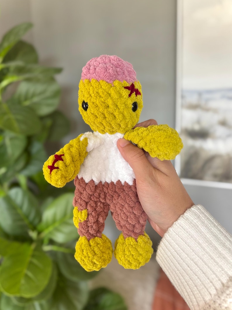 May include: A handmade crochet plush doll featuring a yellow head with a pink top, black safety eyes, and red stitched details. The doll has a white torso, brown legs, and yellow feet, crafted from soft, chunky yarn with a textured, knotted appearance.