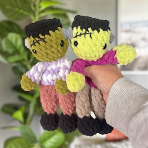 May include: Two handmade crochet plush dolls resembling Frankenstein's monster. One doll has olive green skin, black hair, a white and purple patterned shirt, and dusty rose pants. The other has lime green skin, black hair, a magenta shirt, and tan pants.