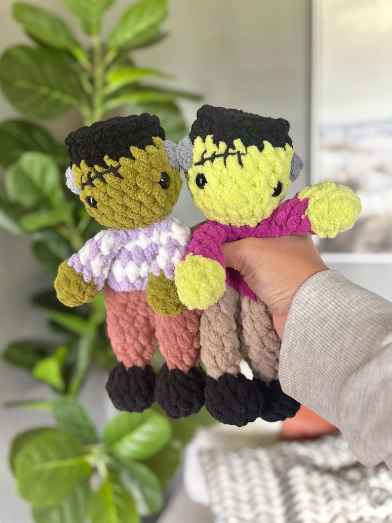 May include: Two handmade crochet plush dolls resembling Frankenstein's monster. One doll has olive green skin, black hair, a white and purple patterned shirt, and dusty rose pants. The other has lime green skin, black hair, a magenta shirt, and tan pants.