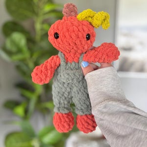 May include: A handmade crochet plush toy shaped like a pumpkin with a round orange head, black plastic eyes, and a small brown stem. The pumpkin wears olive green overalls and has a textured yellow leaf accent attached to the top of its head.