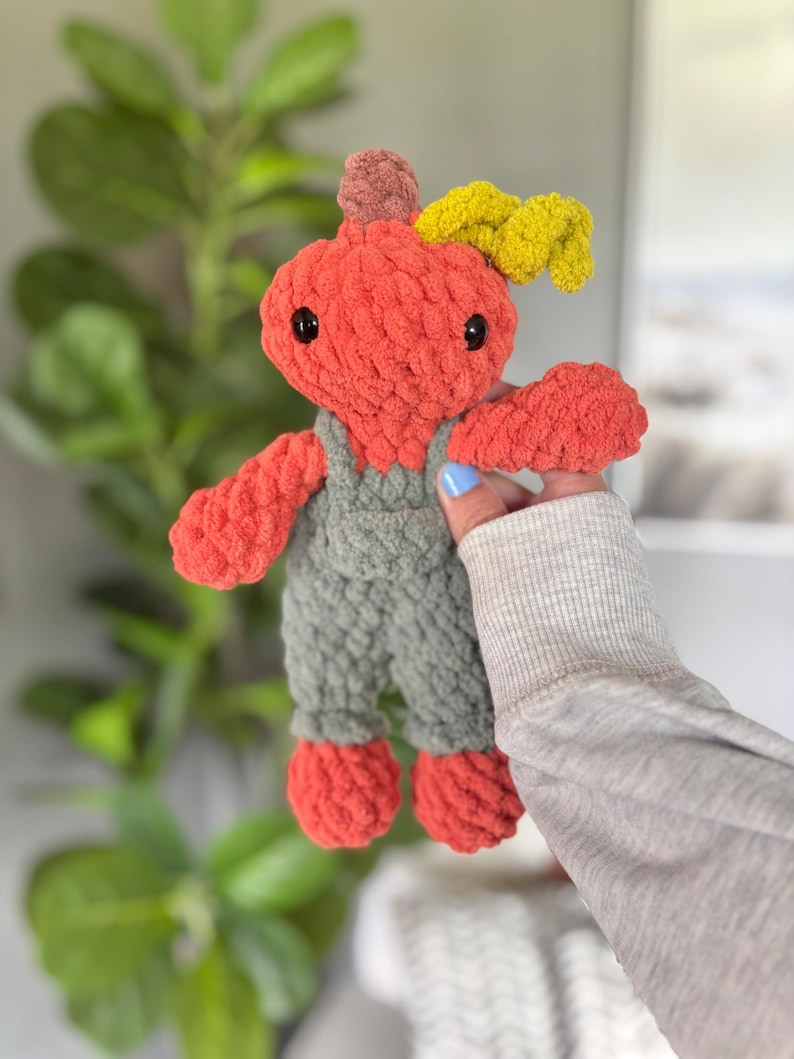 May include: A handmade crochet plush toy shaped like a pumpkin with a round orange head, black plastic eyes, and a small brown stem. The pumpkin wears olive green overalls and has a textured yellow leaf accent attached to the top of its head.